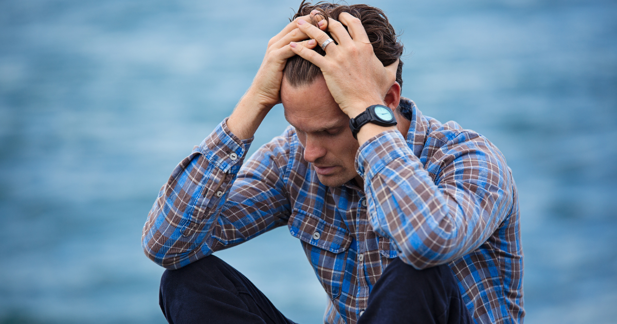 Man with head in his hands and elbows resting on his knees. In the background is and out of focus blue sea.